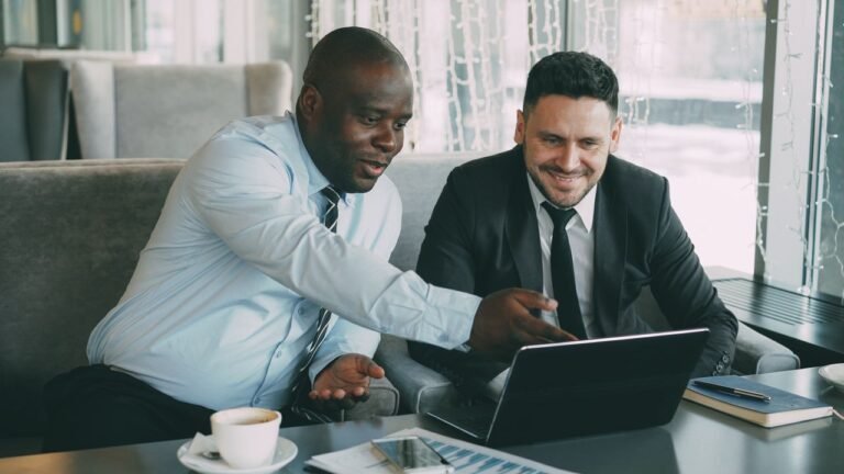 Two businessmen discussing work while using a laptop in a cafe setting.
